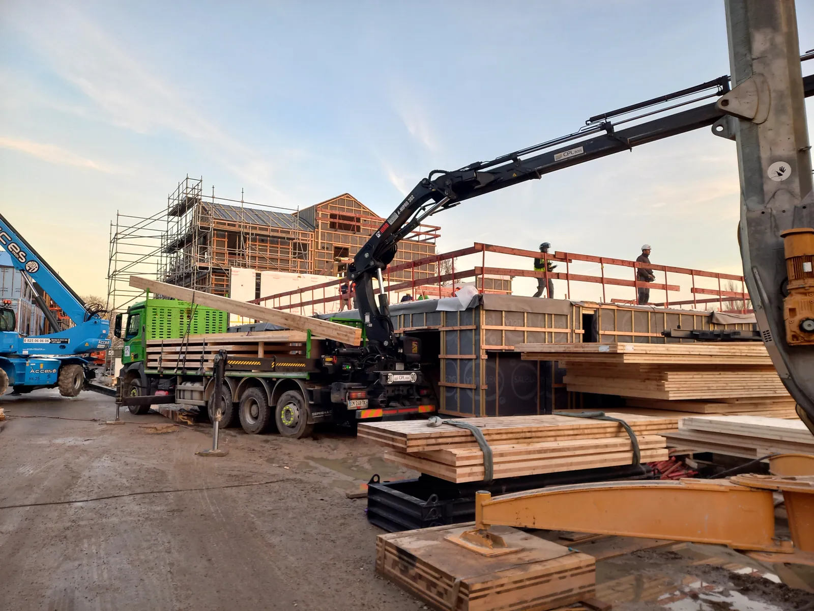 Livraison d'ossature bois sur le chantier de Belle-Île-en-Marne : camion-grue en cours de déchargement, piles de panneaux et de poutres au premier plan, ossatures déjà levées et échafaudages en arrière-plan, en fin de journée.