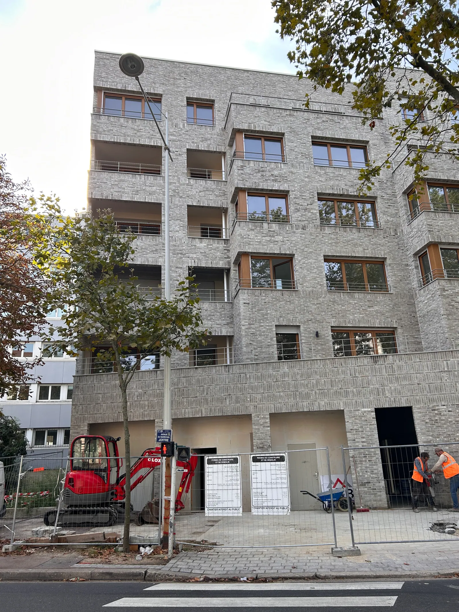 L'immeuble de brique en fin de chantier, vu depuis l'avenue : clôtures de chantier, mini-pelle et panneaux de permis de construire au pied de la façade.