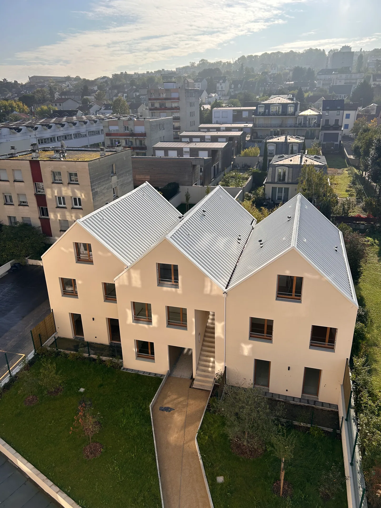 Vue plongeante sur les trois maisons à pignons de Recto-Verso : enduit clair, toitures en bac acier, jardin et cheminement en pied, tissu pavillonnaire de Nanterre à l'arrière-plan.